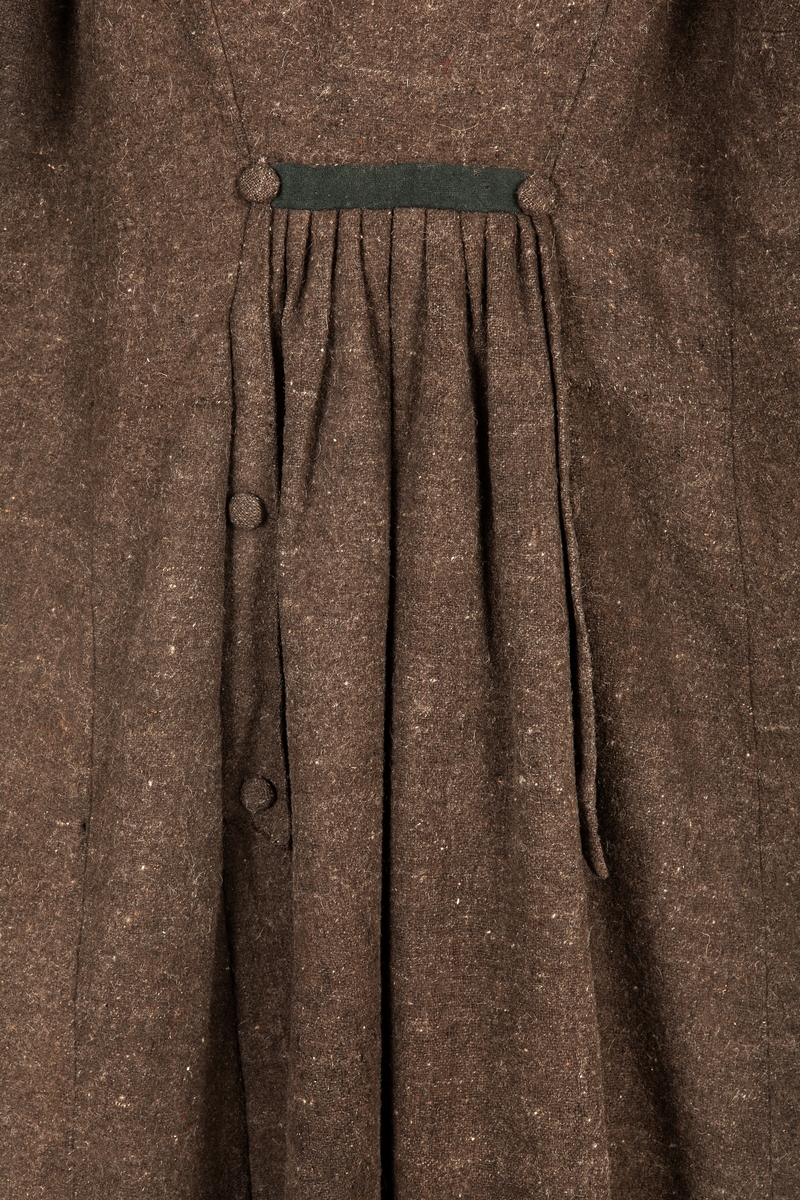 Rock av brungrått tuskaftat ylletyg med inslag av grövre ofärgad ulltråd. Rak krage av mörkgrönt kläde, dubbelvikt (20 cm hög, totalt). Knäppning på framstycket med 10 stycken knappar i dubbla rader, överklädda med rockens tyg. Dessutom knäppning med 4 stycken 10 cm långa slejfar av grönt kläde, som knäpps med gröna tygöverklädda knappar. På ärmarna gröna slag fästade med en knapp. Ryggsidan skuren med rynkning under grön slejf. Rockens insida fodrad med brunvitt kyprat linne med ylleinslag.