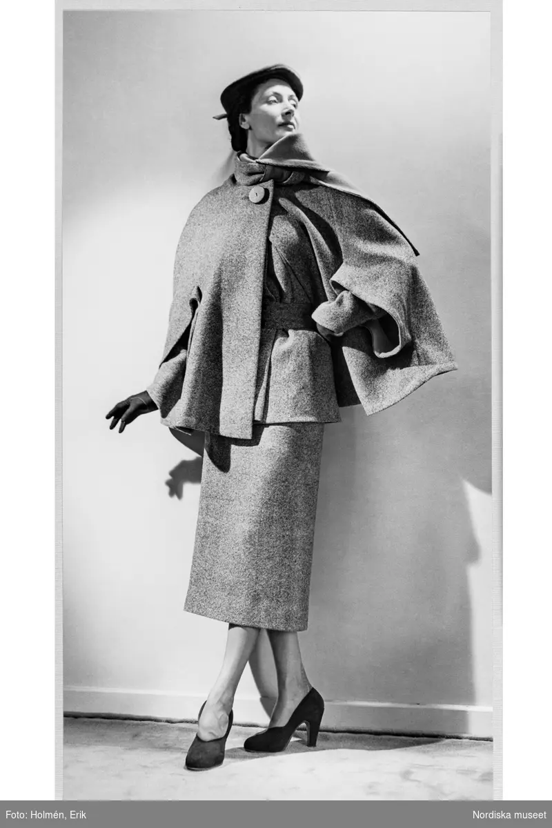 Mannekängen Margareta Berglund gift Nordlund (1946) poserar iklädd ytterplagg i provrummet på NK:s Franska damskrädderi. Stusslång jacka med brett skärp i samma tyg, vadlång pennkjol. Över detta bär hon en stusslång cape med draperad krage och knäppt med en stor knapp. Hatt, handskar och pumps.