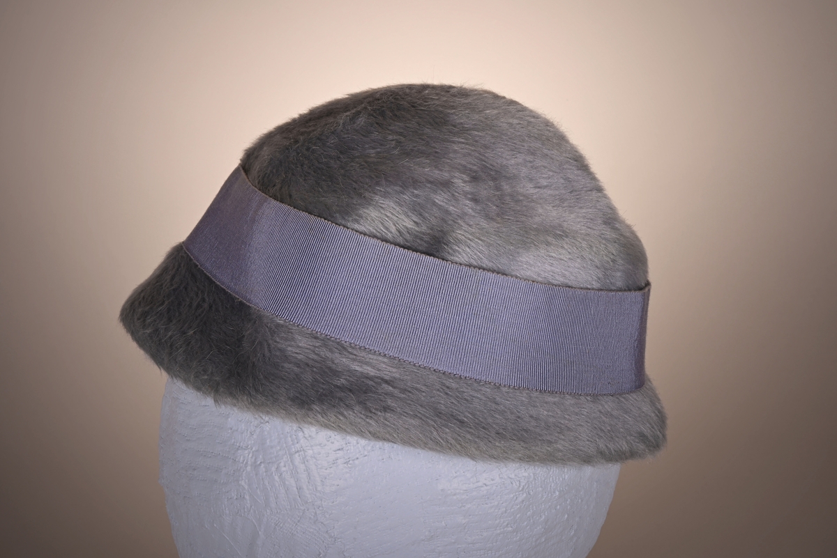 Blågrå damehatt i korthåret fuskepels med smal brem. Materialet er formbart. Rundt hatten er det festet et silkebånd, som er knytt i en sløyfe i bakhodet.