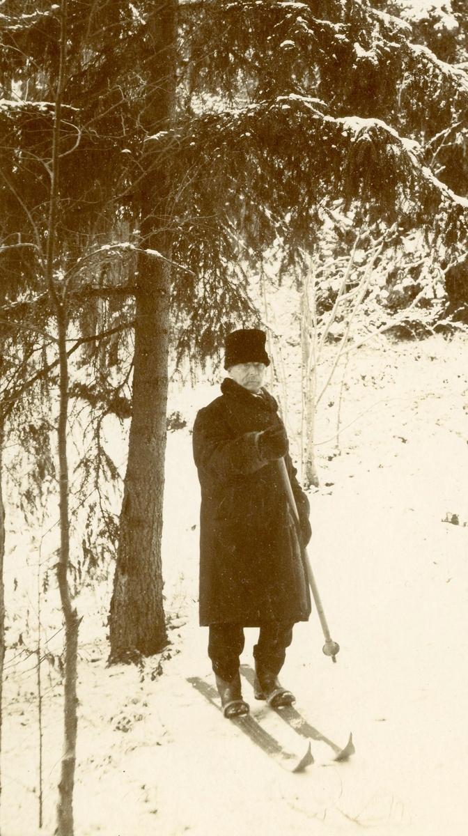 Henrich O.B.Homann på ski, en stav. En mann på ski på toppen av bakken og en til høyre på kne. I vinterskogen.