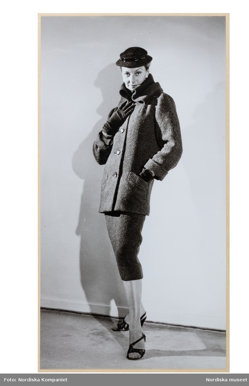 Mannekäng, troligen Margareta Berglund gift Nordlund (1946), visar höstplagg i provrummet vid NK:s Franska damskrädderi. Dräkt av tweed med stusslång rak jacka, knäppning fram, stor ståkrage och snedfickor. Vadlång rak kjol. Hatt, handskar och remsandaletter av slingbackmodell.