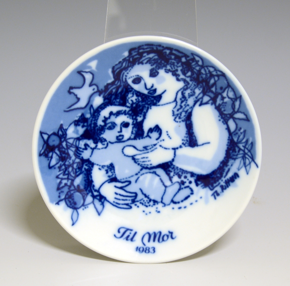 Morsdagsplatte av porselen. Hull til oppheng bak. Hvit glasur. Dekorert i blått med sittende kvinne som mater et lite barn, tekst Mors Dag 1986.
