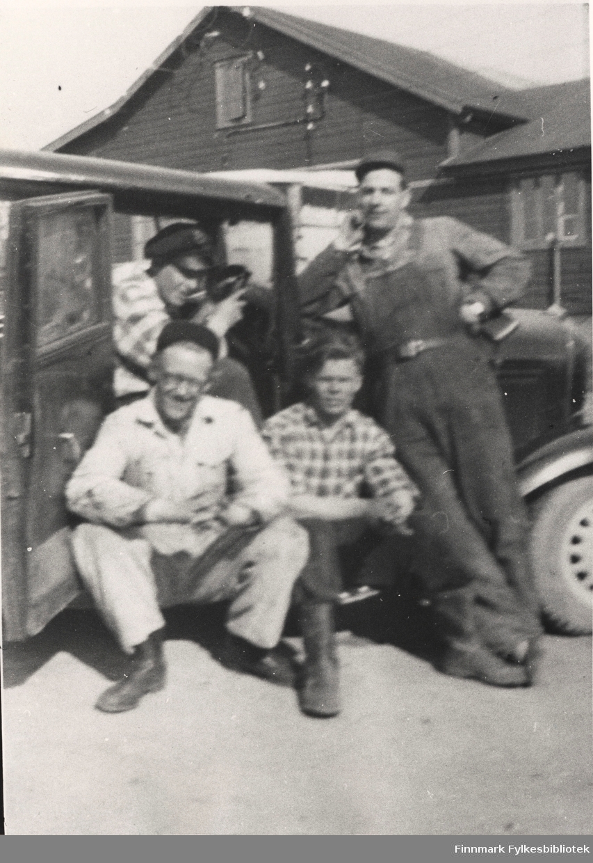 Fire menn ved en bil som kan være en Ford 1932. Foran til venstre sittende på stigbrettet: Lars H. Hofseth og Dikkanen. Bak, sittende i bilen, Lindseth og Erling Kvam stående lent mot panseret. I bakgrunnen er verkstedet i Vegsletta i Vadsø.