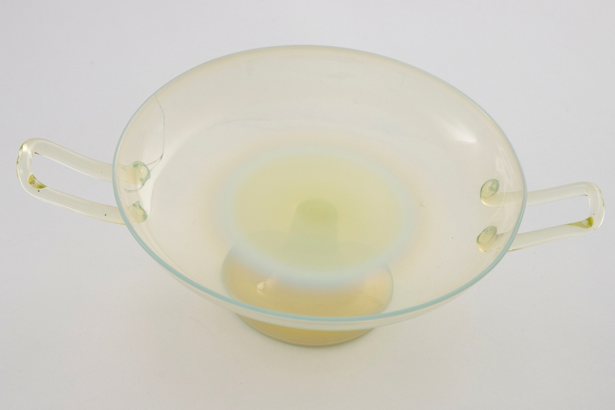 Skål i gultonet glass med opaliserende partier. Lav og vid kupa som bæres av en trumpetformet stett. Påsatte hanker på hver side av kupa.