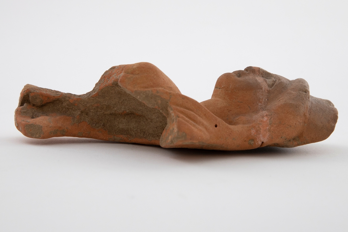 Fragment av figurin i terrakotta, øvre halvdel av kroppen er bevart. Fremstiller en kvinne iført drakt med bølget hår. Figurinen har brudd ved ansiktet og er senere limt. Lyst, rødlig gods.