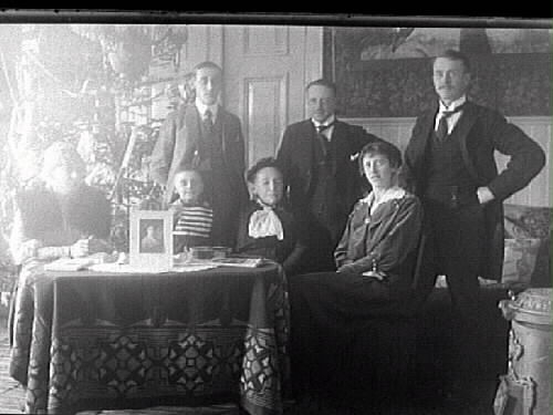 Familjebild vid ett bord klätt i jugendduk och i bakgrunden en julgran. På bordet står ett fotografi.