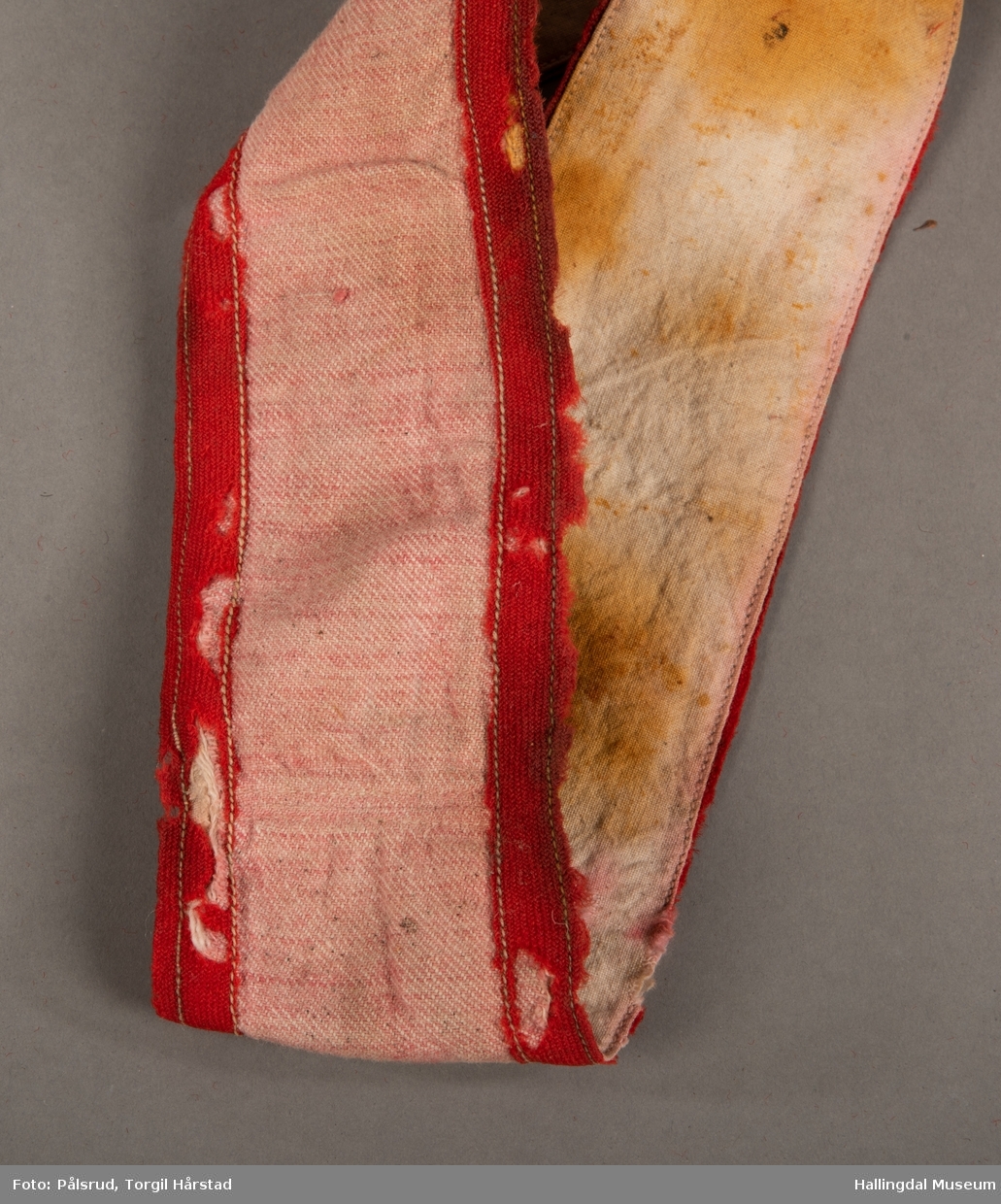 Et langt rødt og hvitt reiveband - å reive = å binde; brukt på spedbarn - med slitt, rød kant, og et slitt, rødt bånt i ene enden som er påsydd for hånd. Reivebandet er laget i et stripet, vevd ullstoff i hvit og lys rød. Er velbrukt og innsiden bærer preg av misfarging av det hvite bomullstoffet.