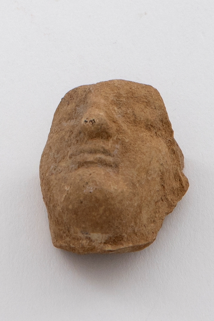 Fragment av figurin i terrakotta. Fremstiller et hode, hvor bare deler av ansiktet er bevart. Grålig gulfarget gods.