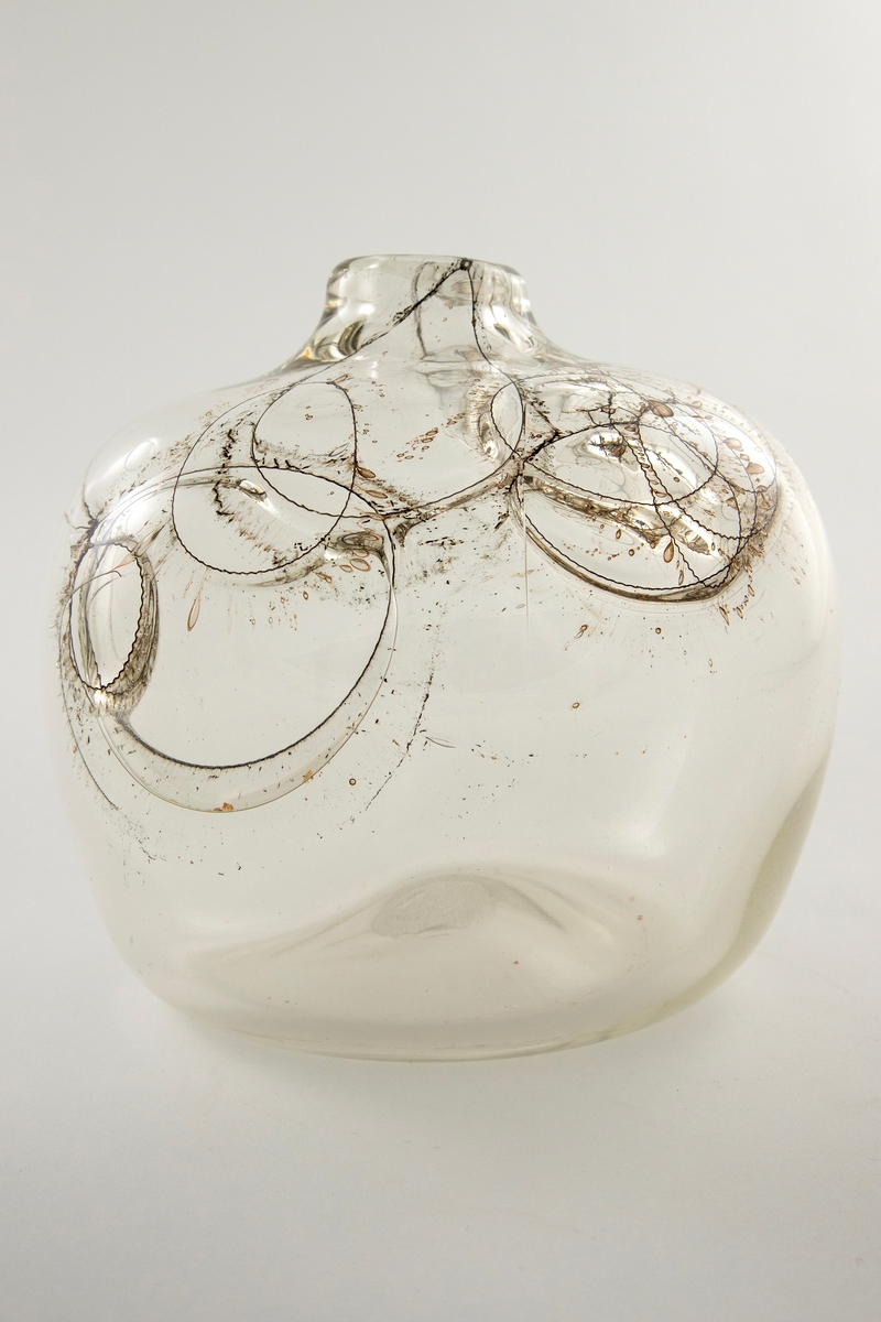 Flattrykket kuleformet vase i klart glass med kort hals. Korpus er dekorert med innlagte ringer av metalltråder på vasens øvre halvdel, og ut ifra disse stråler lange, spisse luftbobler. Puntemerke under vasens bunn.