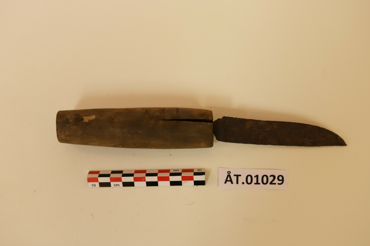 Rustfarget, svart kniv av jern med treskaft. Treskaftet er sprukket på begge sider av knivbladet. 