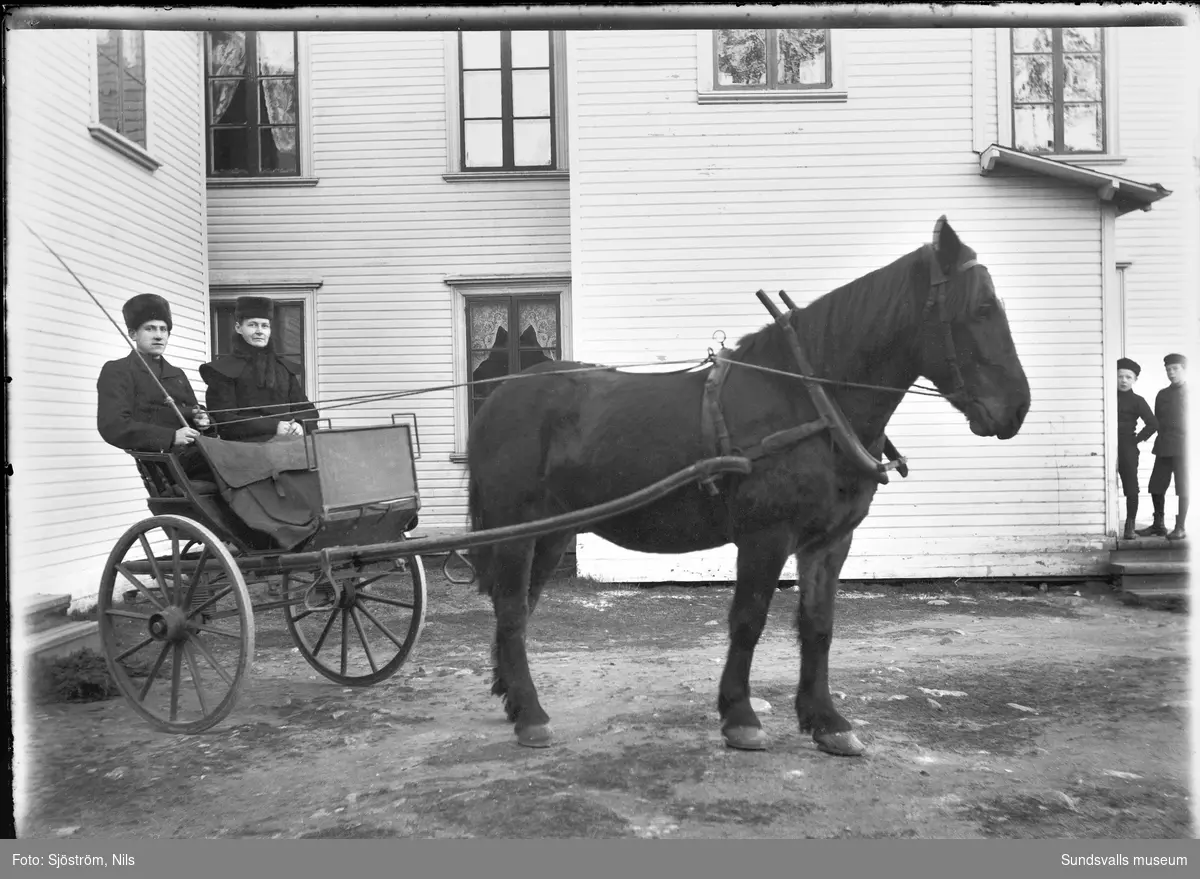 Hästekipage utanför fotograf Nils Sjöströms bostad och ateljé i Vapelnäs. I den tvåhjuliga vagnen sitter en man och en kvinna, båda iklädda pälsmössor. I vänster bildkant syns två pojkar.
