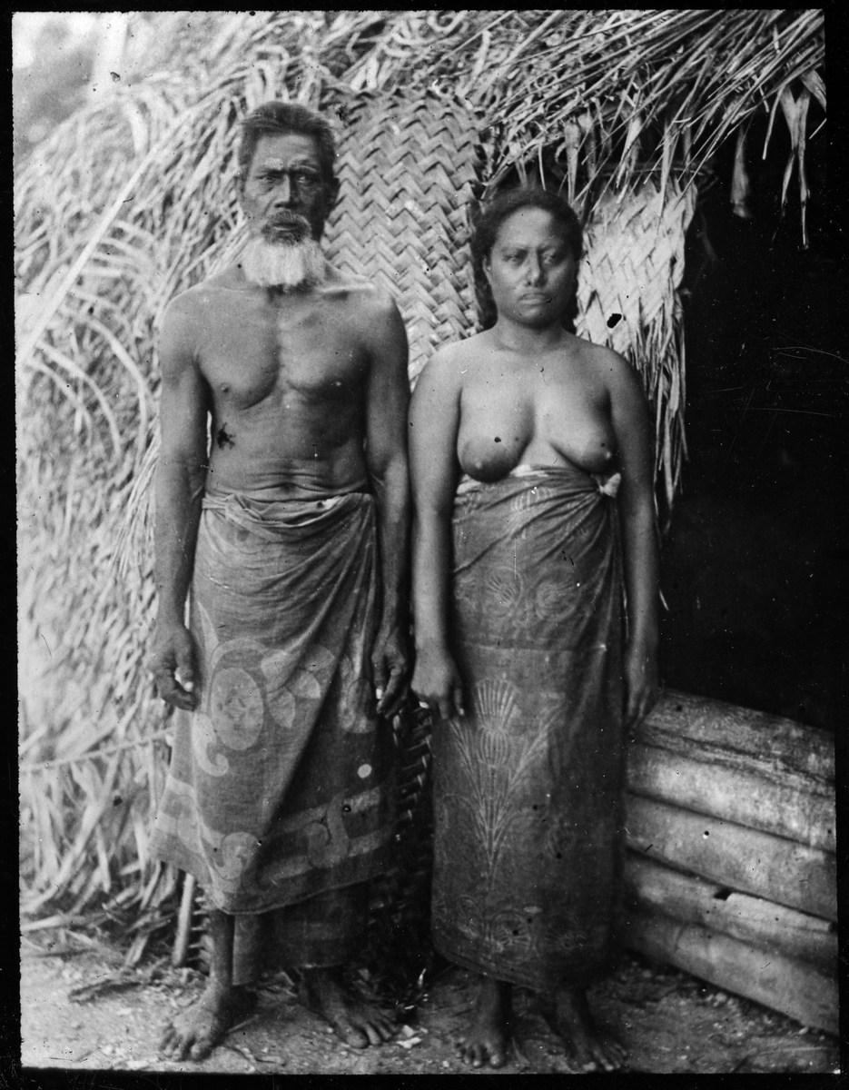 Bilden visar en man och en kvinna från ursprungsbefolkningen på Fakarava klädd i en traditionella dräkter som står framför en hydda. Enlig Etnografiska museets bildinformation är det far och dotter, Tairi och Taroi.