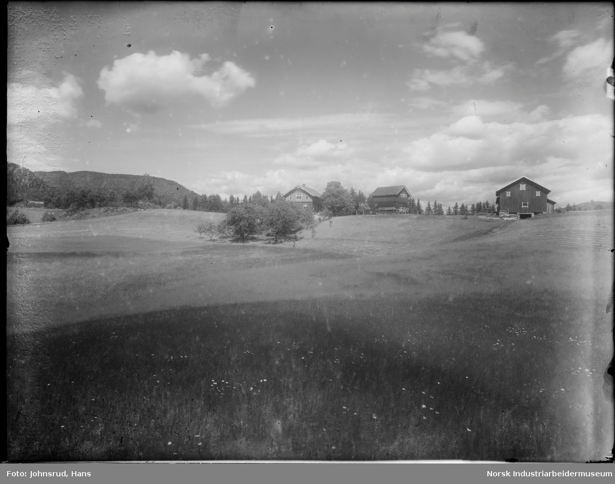 Jorde og eng foran en gård. Et gårdstun med et bolighus, et stabbur og en låve er synlig midt i bildet. Fjell og skog i bakgrunnen.