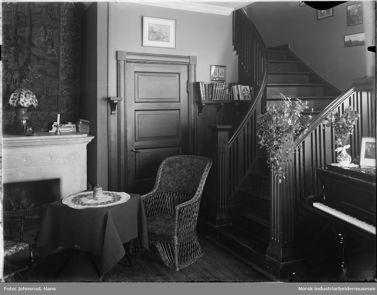 Interiør fra stue. To stoler står rundt et bord foran en dør og en peis i en trappeoppgang. Et piano er synlig til høyre i bildet.