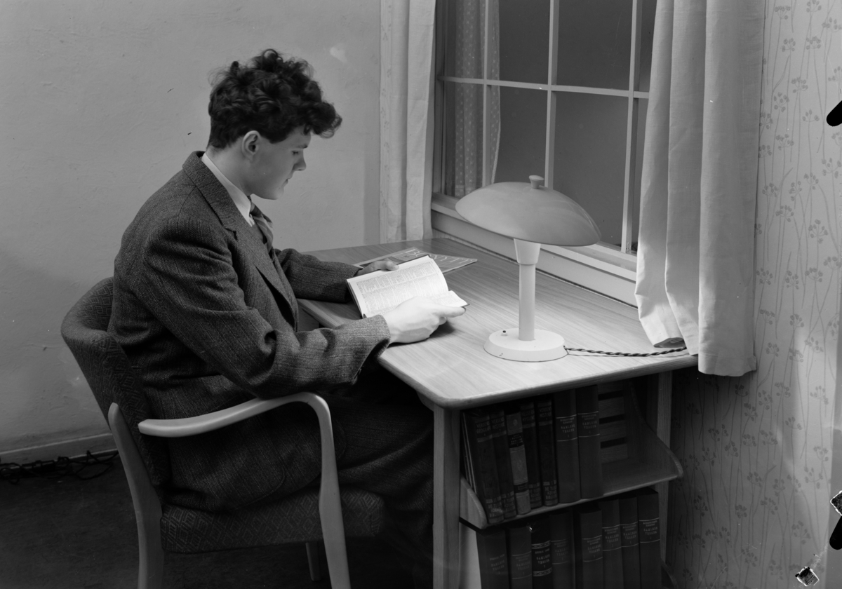 Produktfotografi, P.C.P. bordlampe. En ung mann sitter ved en skrivepult foran et vindu og leser i en bok. Vindusveggen har blomstrete tapet og vinduet har gardiner. Bordlampen på pulten lyser opp det halvmørke rommet. Publisert i Bonytt i februar 1949.