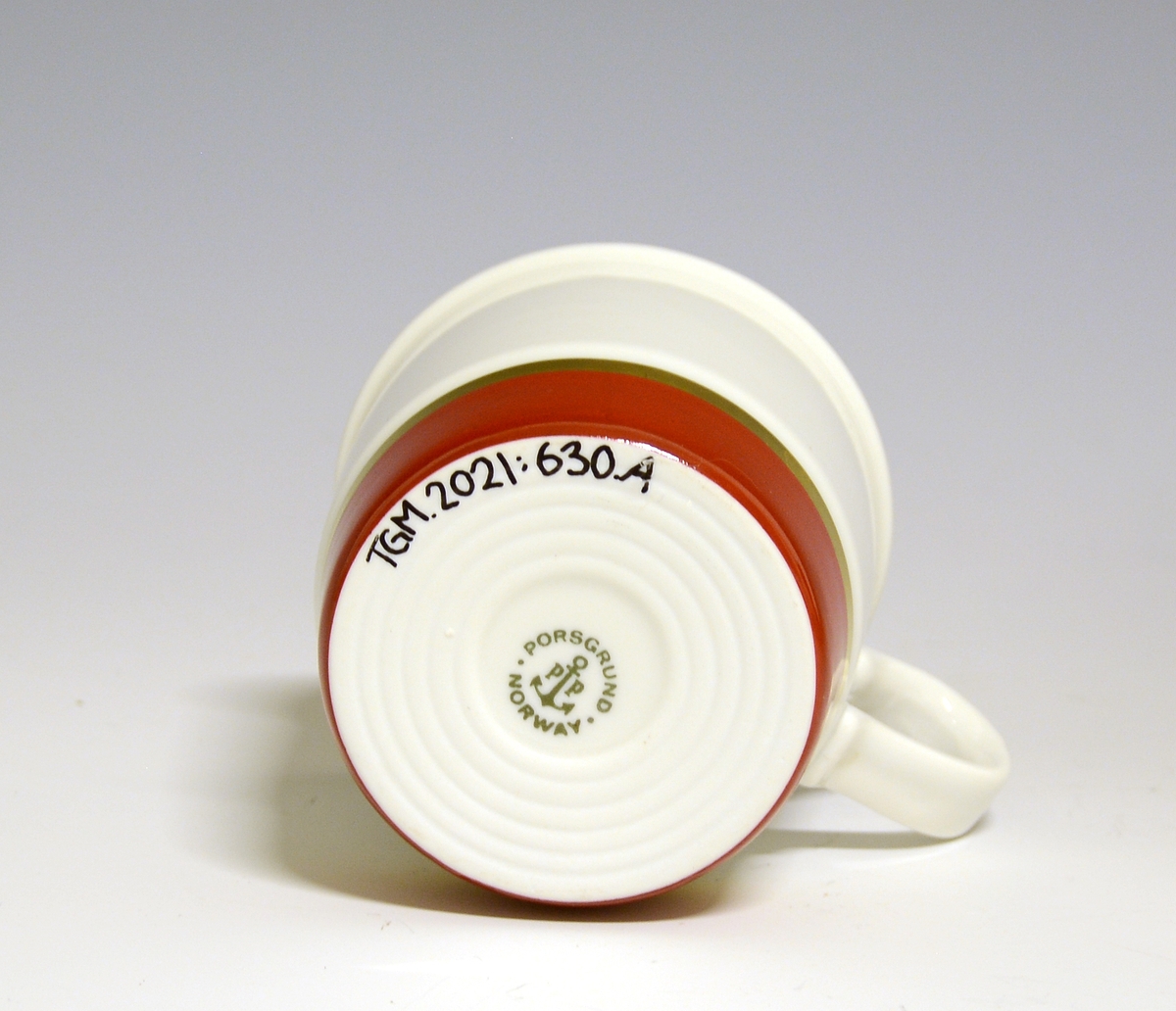 Kaffekopp av porselen. Nederst en bred rød stripe med gullbånd øver. Gullstrek på kantrand.
Modell 444/1 Eystein