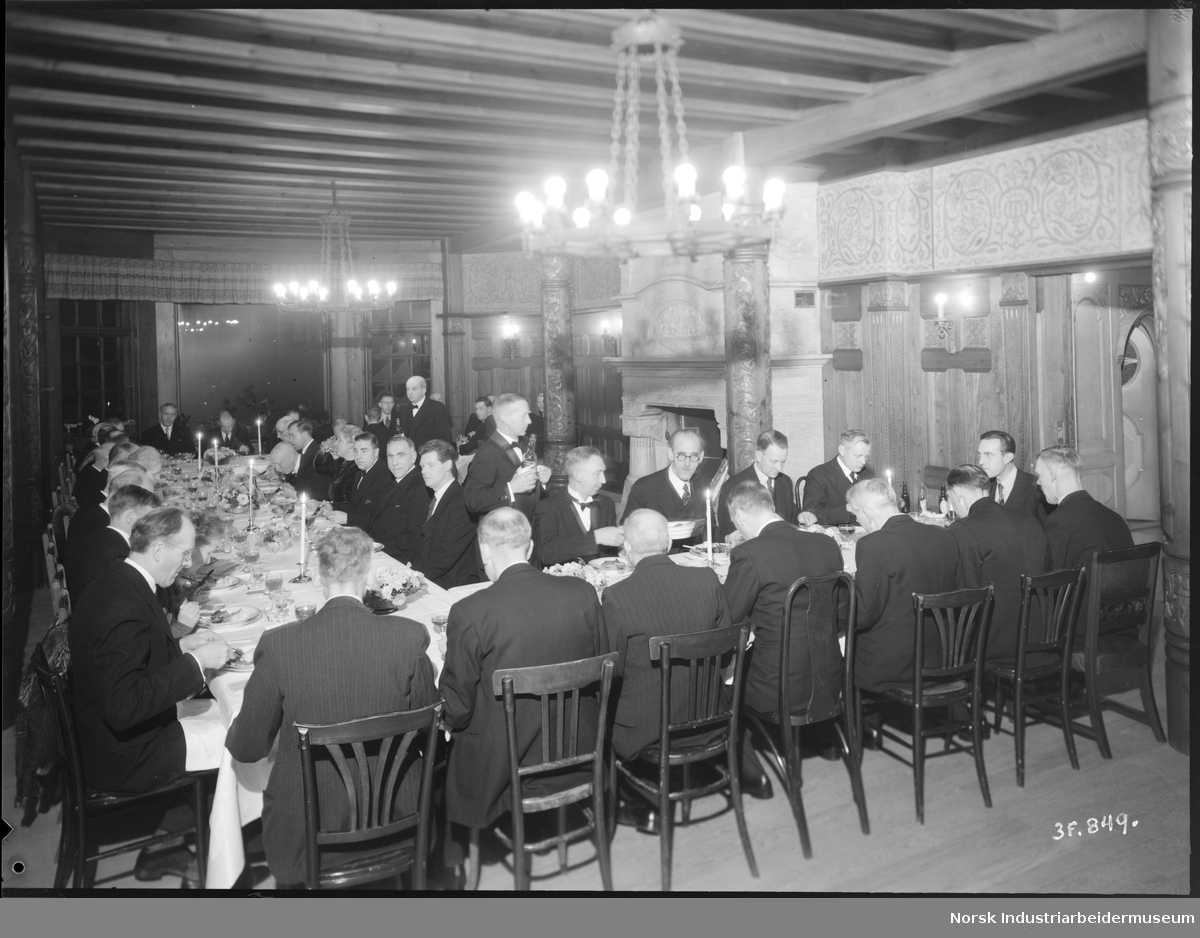 40 og 25 års jubilanter 1945. Jubilantene spiser middag i stuen på Admini. Mannlig servitør serverer drikke ved bordene.