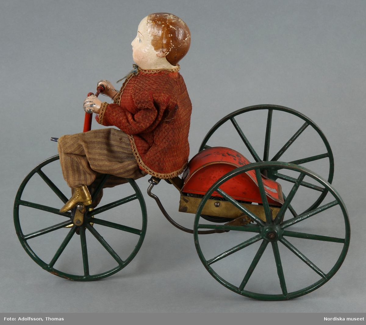 Inventering Sesam 1996-1999:
H 21    L 28 (cm)
Cyklist (a-b), mekanisk leksak, docka med huvud och sköld av papier-mach¿, målade anletsdrag, händer av målad metall samt  skor av mässing. Iklädd närmst röd jacka, beige "skjorta" samt randiga byxor av muslin (1860-tals typ). Kör en trehjulig cykel av metall, lackerad i grönt, blått och rött (a). Nyckel för drivverkets uppdragning (b) (L 6,5 cm).
Möjligen "Stevens & Brown Manufacturing Co." (Amerika), jmf. inv.nr 255.530 samt 299.973.
Charlotta Dobson Hoffman jan 1998