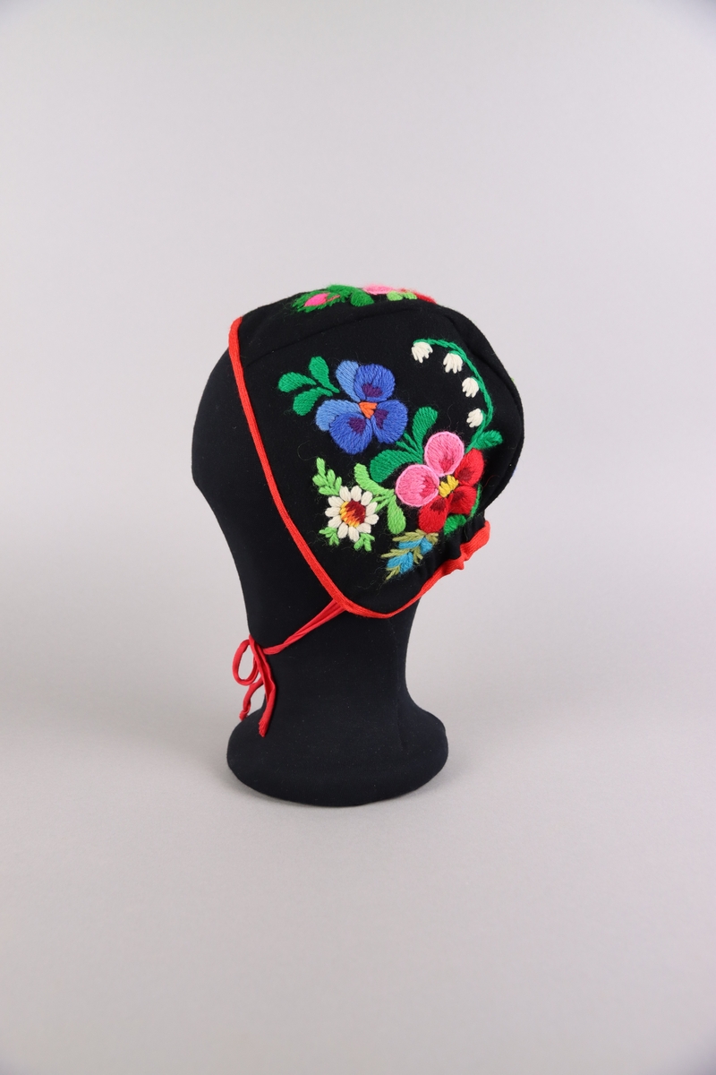 Hätta till flicka av svart kläde med röda band. Hättan är broderad med blomstermotiv av ullgarn i flera färger med påsöm i Dala-Floda-stil.
