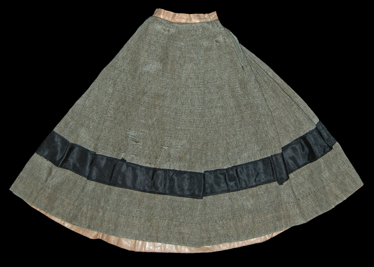Kjol, grågrön med svart band påsytt på nedre delen av kjolen. Beigt band i midjan. Svart sammetsväska med band fastsydd i linningen på kjolen, innehållande en liten spetsnäsduk.