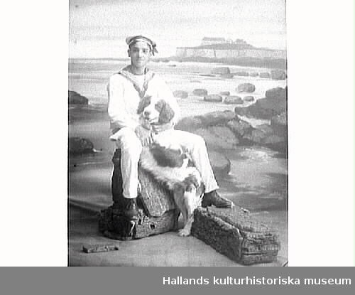 Ung man i sjömansdräkt med hund i famnen. Fond och rekvisita ska ge en illusion av att han sitter på en sten vid havet med Varbergs fästning i bakgrunden.