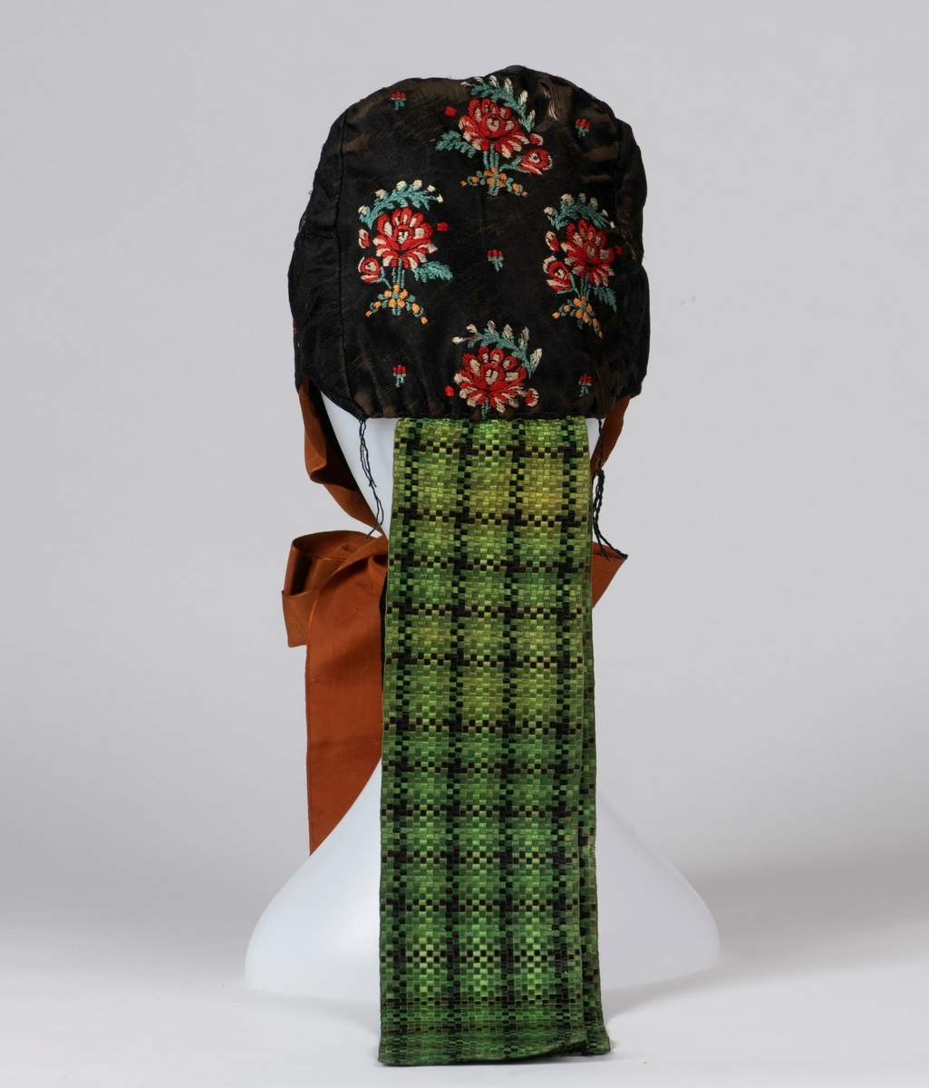 Lue. Kvinnelue, sydd av sort silke brodert med blomstermotiver. Brune knyttebånd av silke, samt nakkebånd vevd med ruter i sort og grønt. 