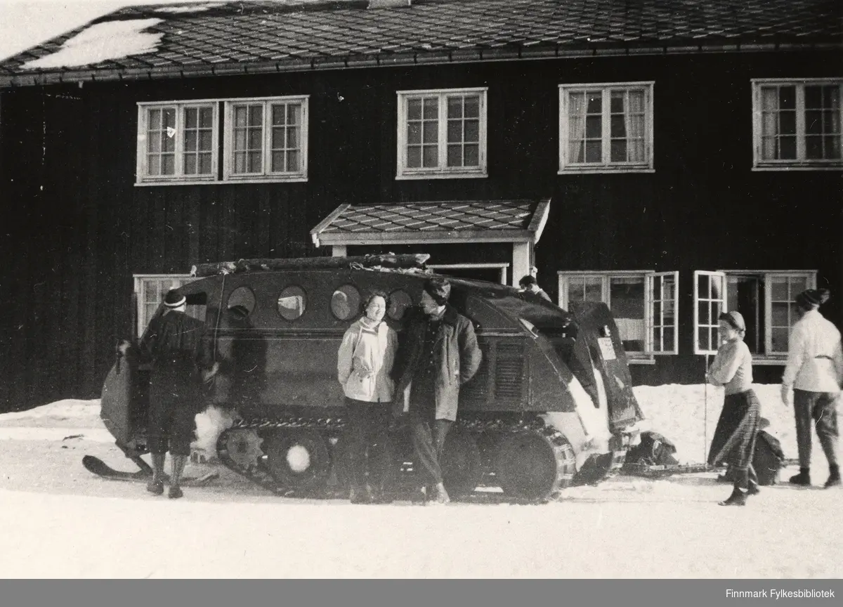 Bilde av en gruppe mennesker og en snowmobil utenfor Kautokeino fjellstue. Fra 1951 til rundt 1966 gikk det på vinteren snowmobilruter fra Kautokeino til Alta, Karasjok og Enontekiö i den finske siden.