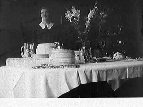 Bild 1: Dukning. Matsalbord med blomsteruppsättning och porslin. En kvinna med vitt förkläde står vid bordet. Bild 2: Närbild av kvinnans ansikte.