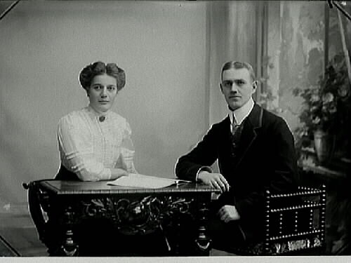 Syskonparet herr och fröken Johansson sitter vid ett bord. På bordet ligger en uppslagen bok.