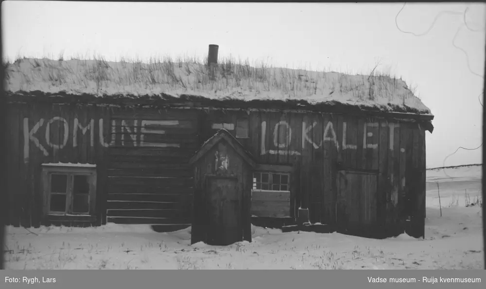Et trehus med gress og torv på taket. På langveggen står det skrevet, med hvite bokstaver KOMUNE  - LOKALE.  Det ligger snø på taket og bakken. Bildet har tilhørt Lars Rygh som var med på minerydding, i Varangerfjorden mellom 1917 - 1918.