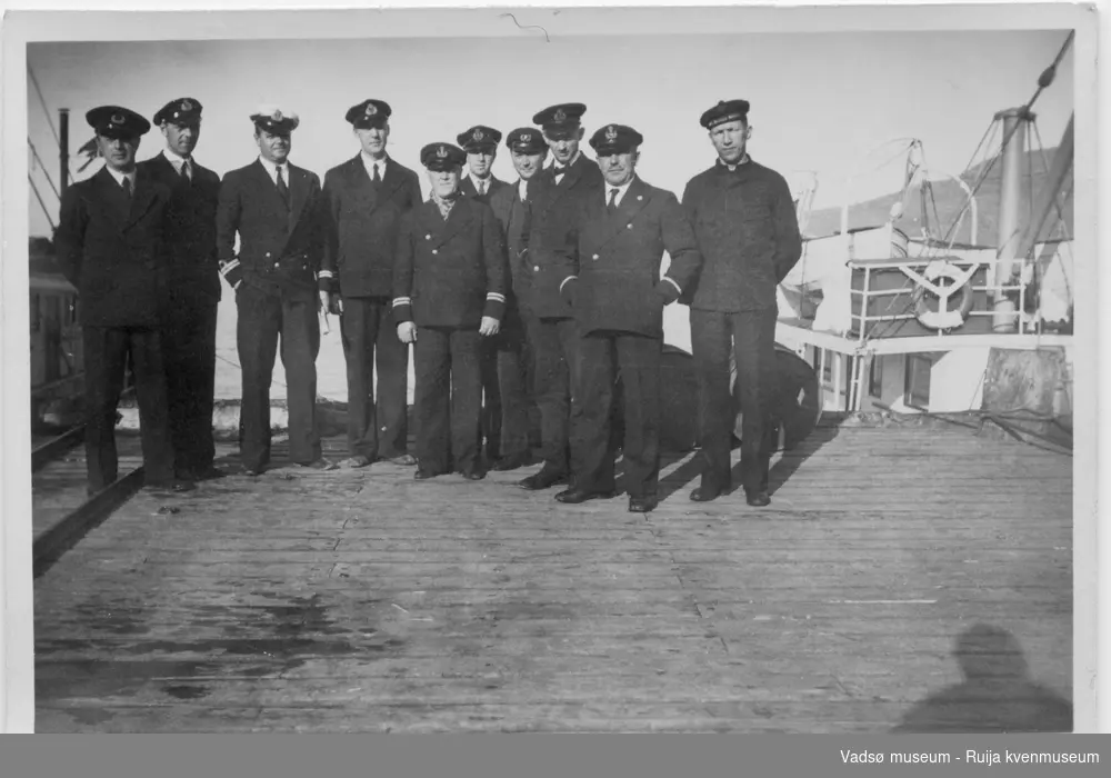 Gruppebilde av ti sjømenn i ulike marineuniformer. De står på en trekai, hvor det ligger båter fortøyd. Bildet har tilhørt Lars Rygh fra Hønefoss. Han var med på mineryddingsoppdrag i Varangerfjorden 1917 - 1918.