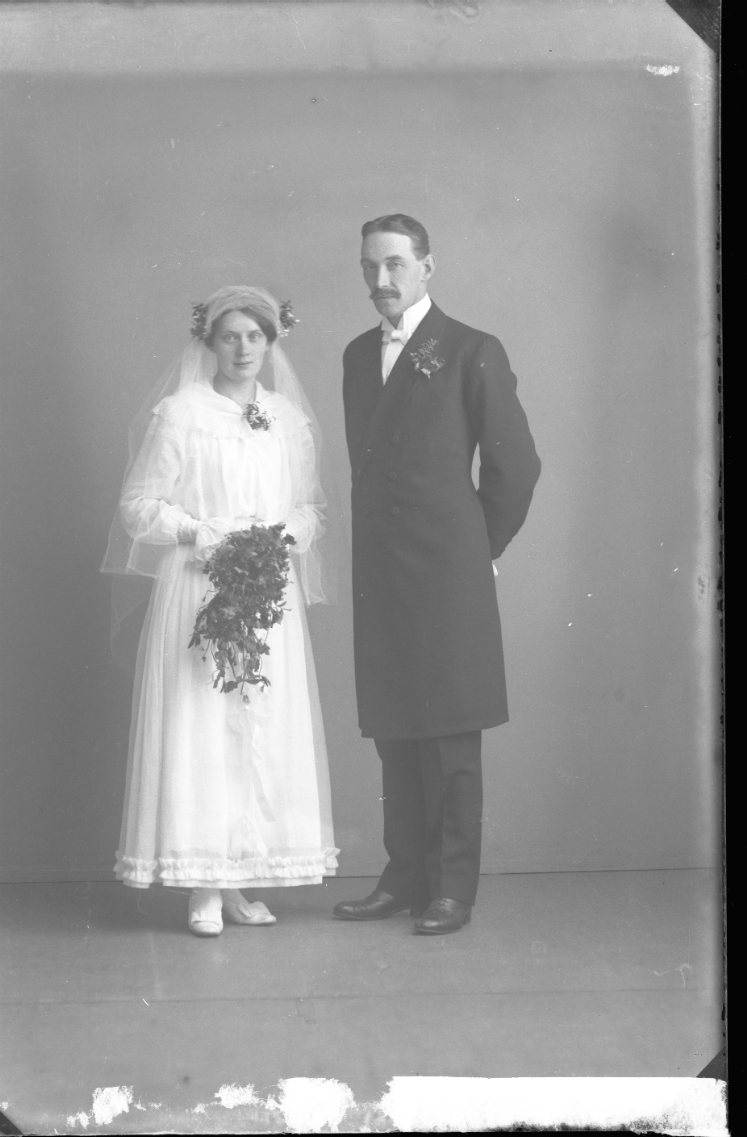 Ett brudpar. Bruden är klädd i vitt och håller en bukett i händerna. Brudgummen har händerna bakom ryggen.