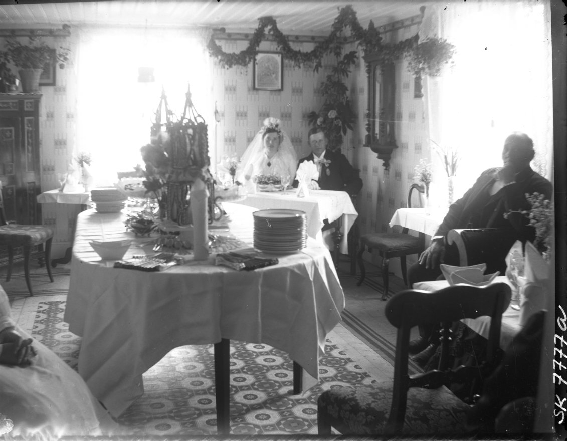 Hemmiljö med ett brudpar längst bort i rummet under en girlang bakom ett bord med vit duk. I framkant står ett bord med vit duk och staplade tallrikar bland annat. En man sitter vid väggen till höger och till vänster anas en kvinna.