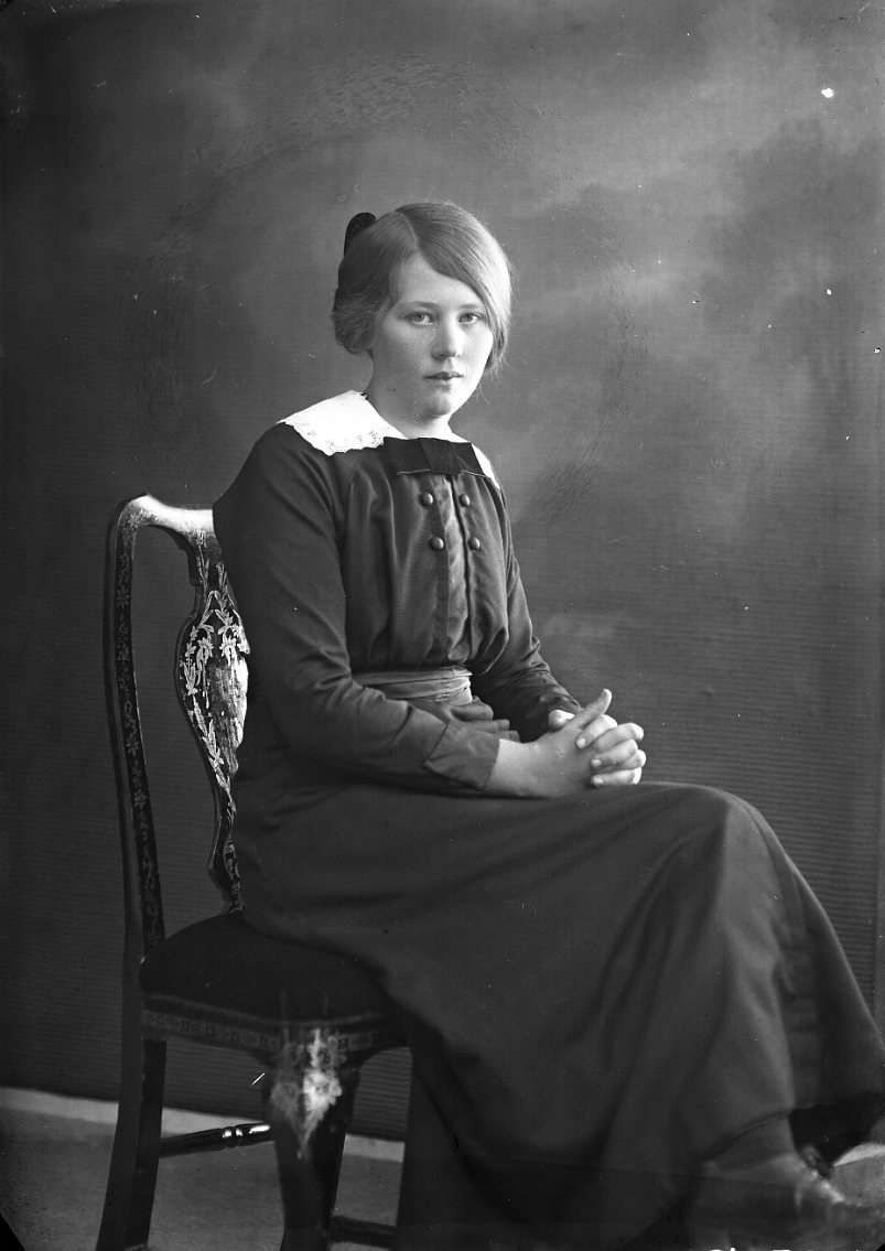Porträtt av en flicka, Ester Johansson från Fallet. Hon sitter på en stol i 3/4-profil vänd mot fotografen med händerna knäppta i knät.