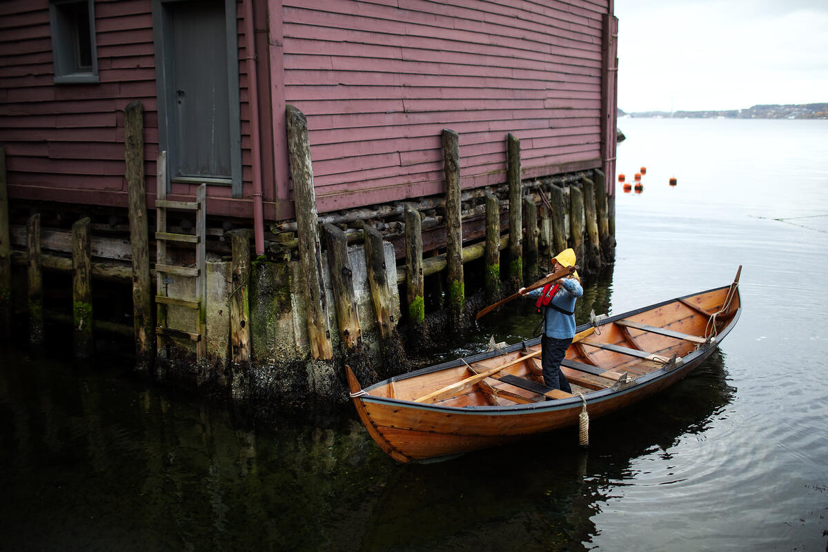 En dame står i robåten mens hun bruker den ene åren til å stake seg vei i den smale kanalene mellom to av sjøbodene.