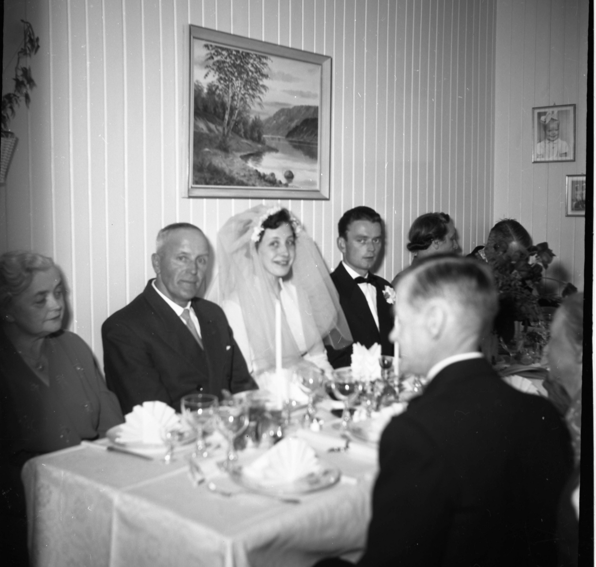 Bilde av penkledde gjester sittende til bords. Bryllupsfest. Brudeparet sees i midten av bildet.