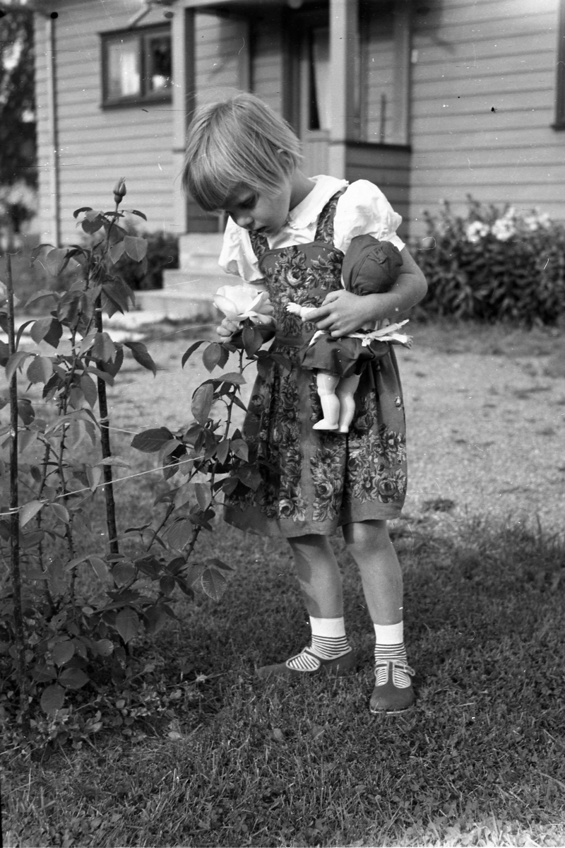 Liten jente i helfigur står og ser på en rose. Hun bærer også en dukke på armen.