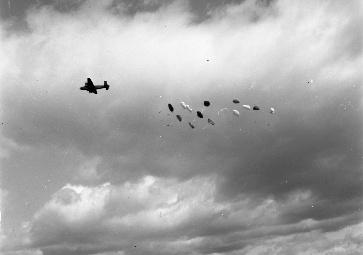 Et bombefly ved navn Consolidated B-24 Liberator slipper ut innhold fra flyet. En rekke fallskjermer er spred utover himmelen.