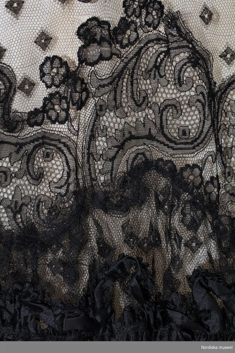 Tvådelad klänning monterad på docka. NK:s Franska damskrädderi.
Tvådelad, korsetterad klänning av vit taft överdragen av svart tyll och spetsar. Klänningen bars av Lydia Maria Bring född Widman (1855-1939) vid en fest på Gripsholm 1904.