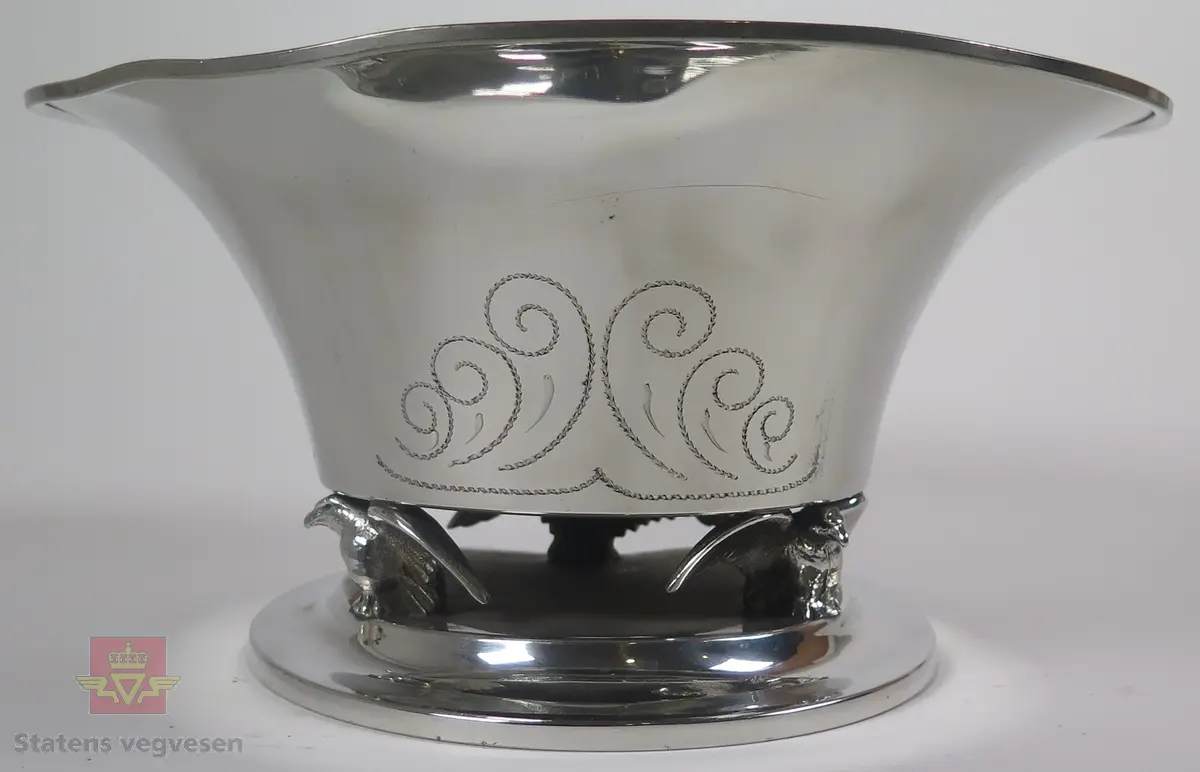 Pokal i sølv formet som en skål/bolle. Det er utformet tre ørner mellom bunnen og toppen av pokalen.