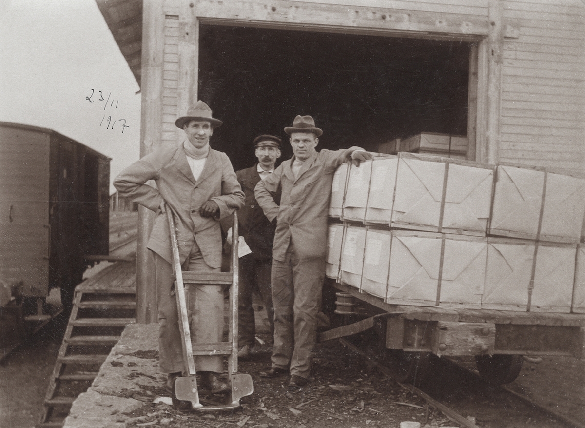 Tre män poserar vid en godsvagn utanför ett magasin, 23/11 1917.
Växjö (?).