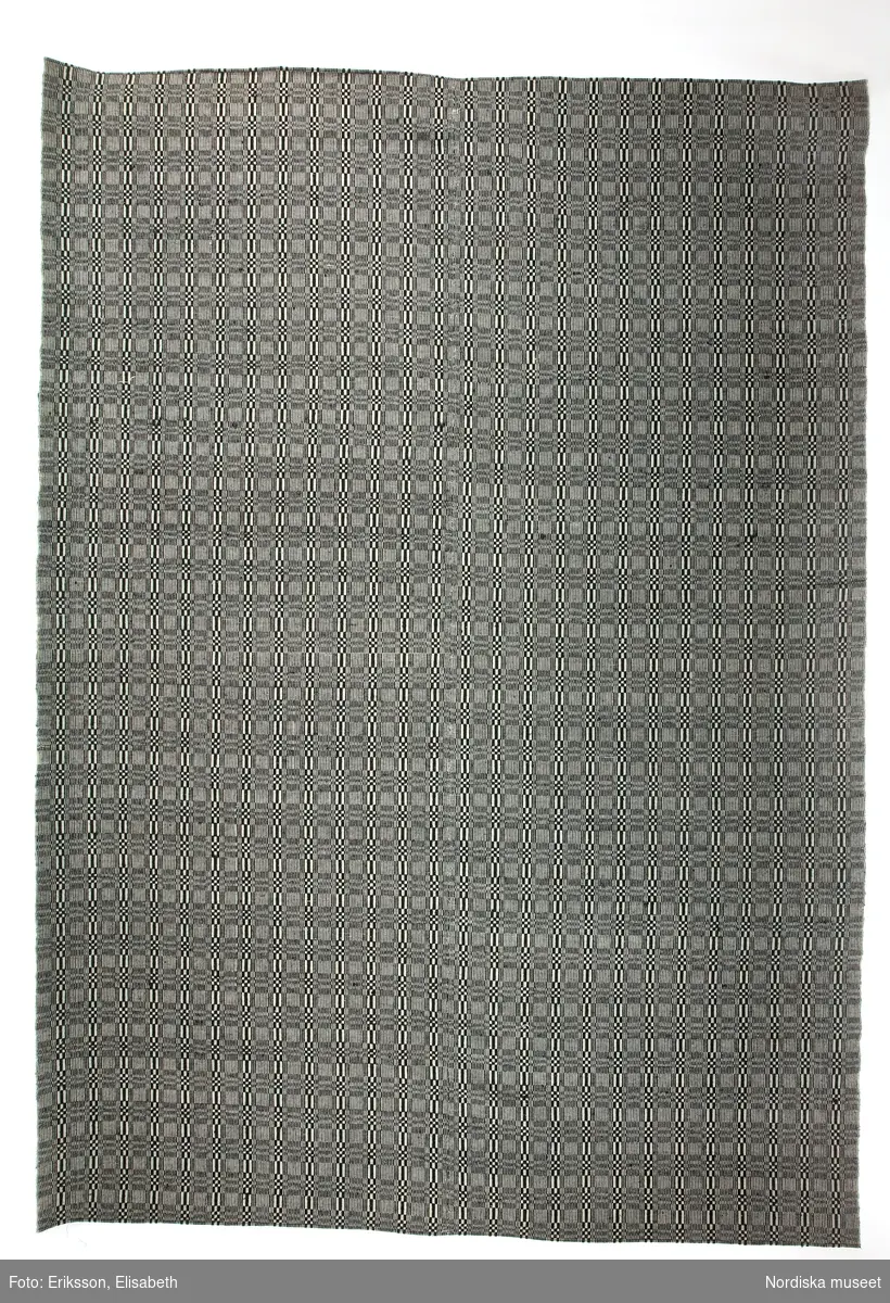 Täcke, varp av z-tvinnat vitt bomullsgarn, 44 trådar på 2 cm, inslag i botten av 2 trådar z-tvinnat grönt bomullsgarn och i mönstret 1-tr. s-spunnet svart ullgarn, förenklad dräll i rutmönster, vävt i 2 våder hopsytt på mitten för hand, maskinsydda fållar.

"Senast använt i en kyrkstuga i Nordmaling" ( 1922)
Linneband med mässingsringar ditsydda av museet för utställning.
Berit Eldvik april 2005