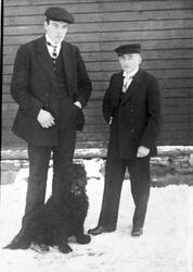 Portrett av to pent kledde menn og en hund stående ved en hu