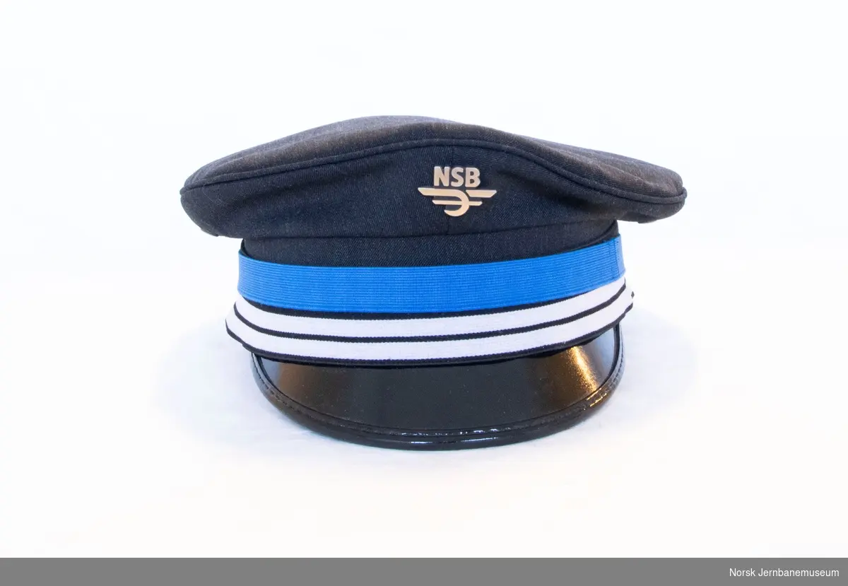 A black peaked cap used by the National state railway (NSB) conductors up until the NSB changed its name to Vy. It has a thick blue stripe and two smaller white stripes, as well as an NSB logo in metal. This spesific cap was used by a female conductor, and it was used in the time period 2013-2019 before she gave it to the museum in 2020. 