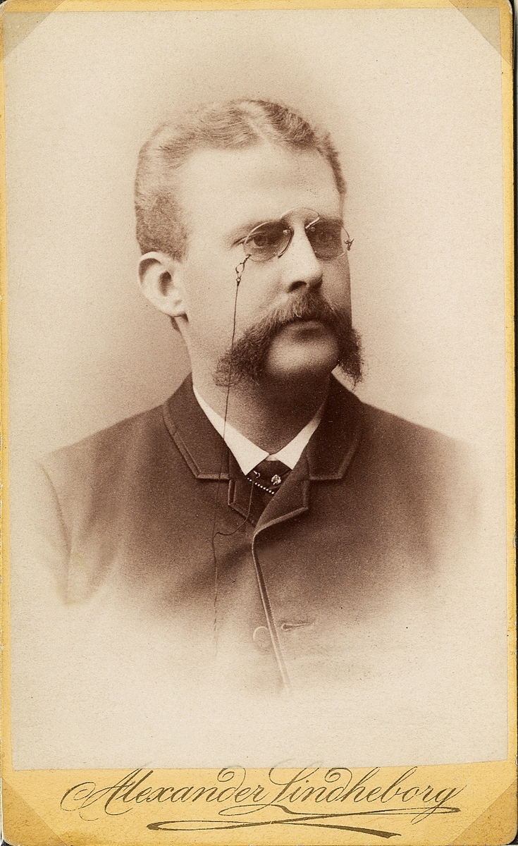 Porträttfoto av en man med mustascher och pincené, klädd i kavajkostym med stärkkrage och slips. 
Bröstbild, halvprofil. Ateljéfoto.