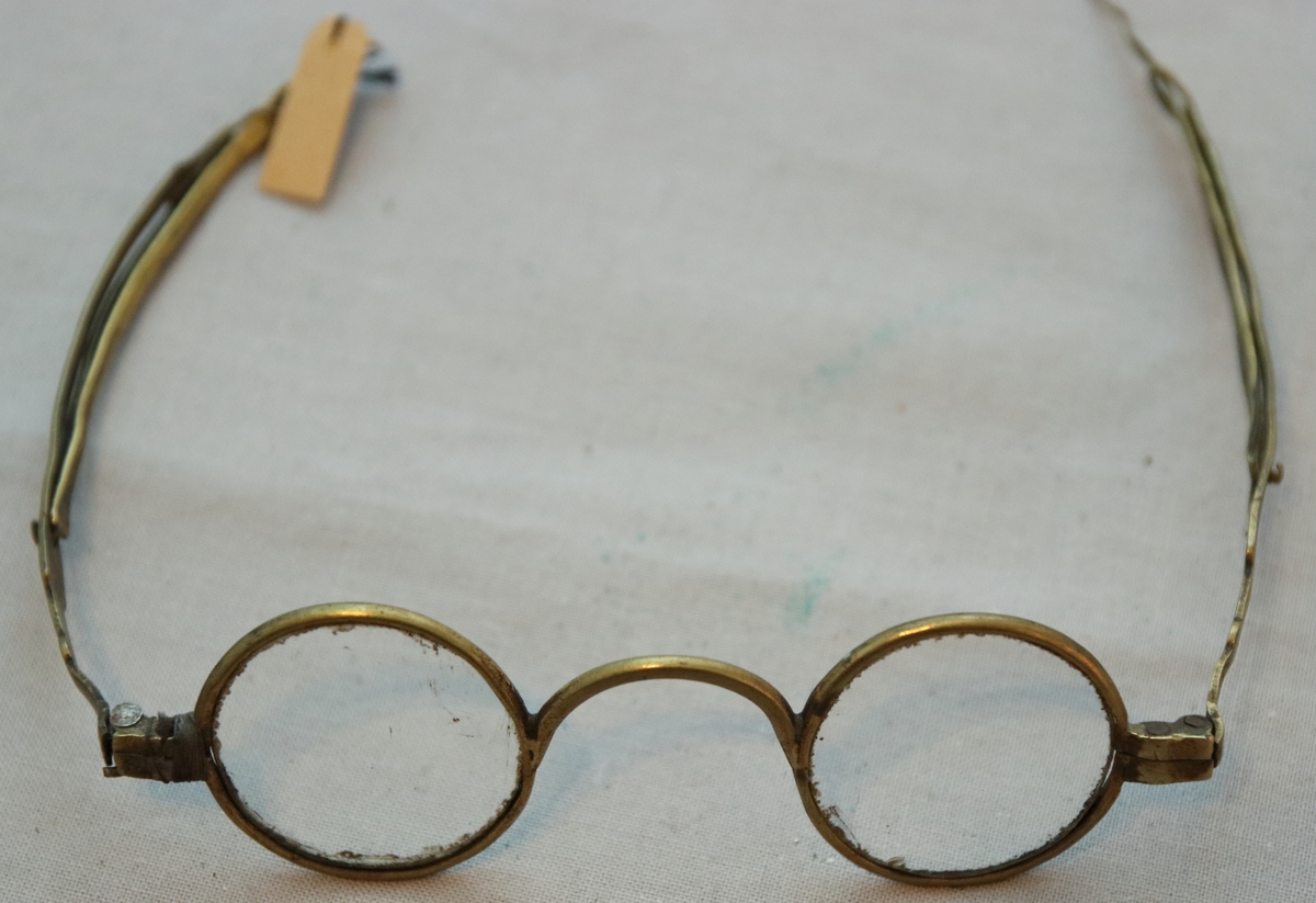 Glasögon med cirkelrunda glas. Ramar och skalmar av mässing, de senare med skjutbar förlängning, försedd med ögla.