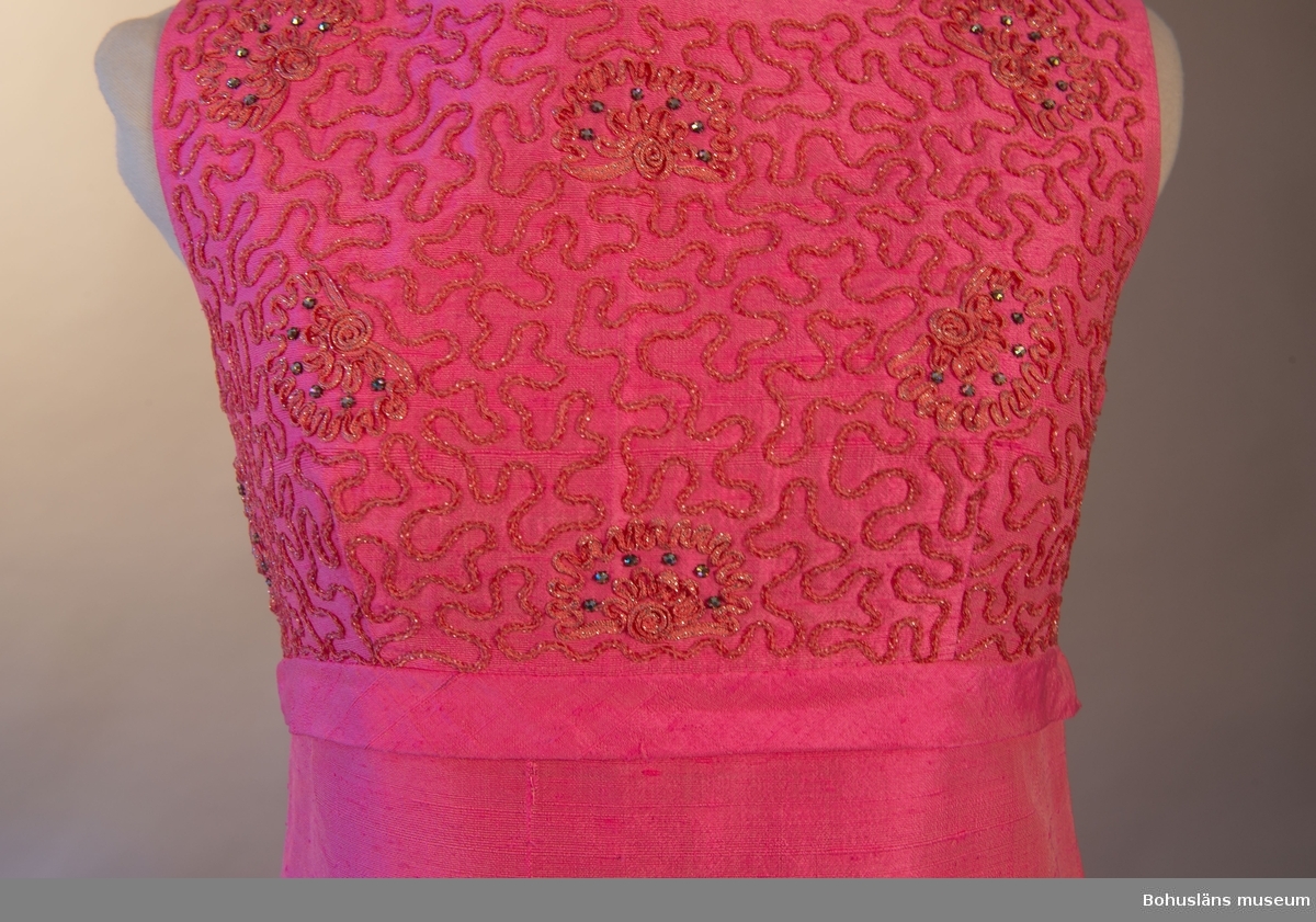 Rosa långklänning band något över midjan som löper runt klänningen. Över bandet, på klänningens framsida, mönstrat tyg ornamenterat med musslor i oregelbundet mönster. Baksidan har ett släp fastsytt i bandet.
