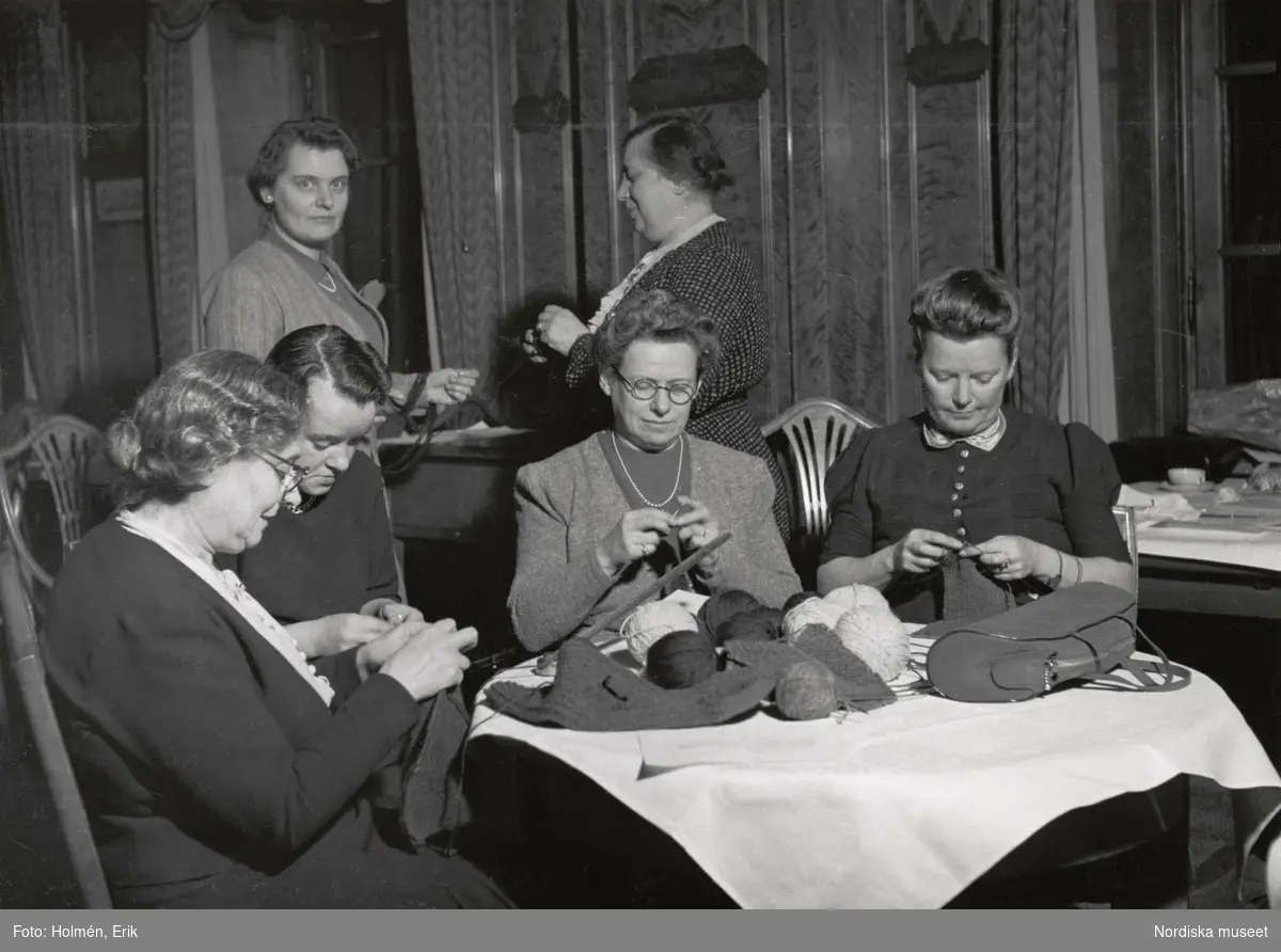 Beredskapsstickning på Nordiska kompaniet. Fyra kvinnor sitter runt bord och stickar, två kvinnor står bakom. 