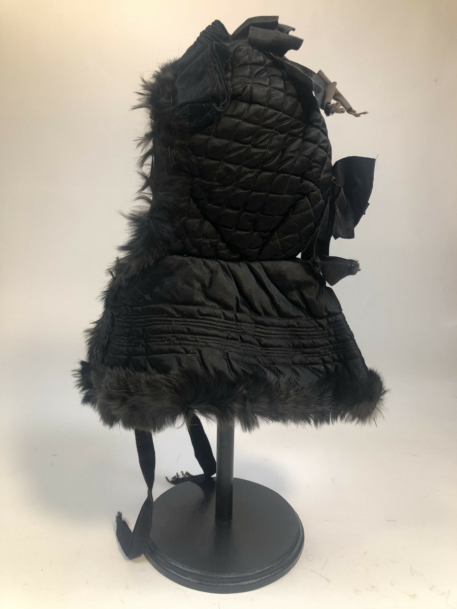 Reisekyse, såkalt "kråke", av glatt, svart stoff, kanskje blanding av ull og silke. Kysa er vattert og dekorert med pelskant og silkeband. Den knytes med svarte band foran.