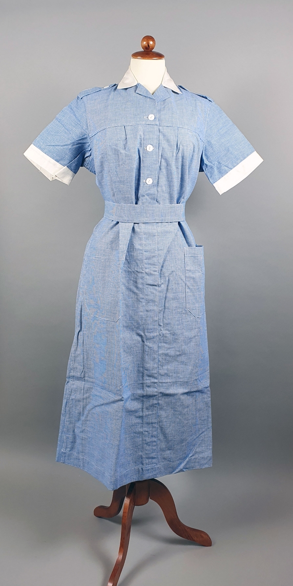 Blå bomullskjole med korte ermer, påsydde lommer og belte med knapp. Hvite knapper foran og spensel på skuldrene. Hvit skjortekrage.